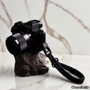 May include: A black Sony camera with a lens and a black wrist strap rests on a gray rock. The camera is positioned against a white and gold marble background. The wrist strap is made of woven black material.