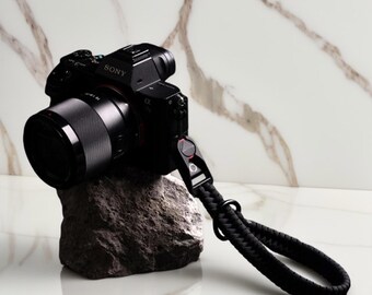 May include: A black Sony camera with a lens and a black wrist strap rests on a gray rock. The camera is positioned against a white and gold marble background. The wrist strap is made of woven black material.