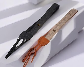 Könnte beinhalten: Zwei Kameragurte, einer in Schwarz und der andere in Beige, werden gezeigt. Der schwarze Gurt hat eine dunkelgraue Webstruktur, während der beige Gurt eine hellere Webstruktur aufweist. Beide Gurte haben Lederakzente und Metallbeschläge.