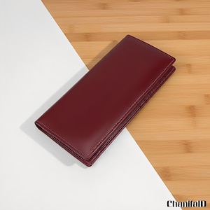 Peut inclure: Un portefeuille en cuir bordeaux, de forme rectangulaire, est posé sur une surface blanche et imitation bois. Le portefeuille est fermé, présentant une texture lisse et polie avec des bords cousus. Le design est épuré et minimaliste.