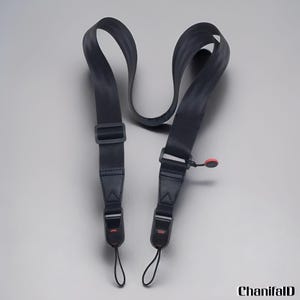 May include: A black camera strap with adjustable buckles and a red accent. The strap is made of a durable, woven material and features a loop for attaching to a camera. The strap is designed to be worn around the neck or shoulder.