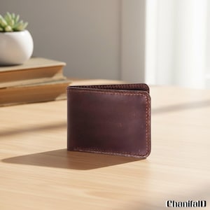 May include: A brown leather wallet with a bi-fold design sits on a wooden surface. The wallet is a rich, dark brown color with visible stitching around the edges. A small succulent plant in a white pot and a stack of books are in the background.