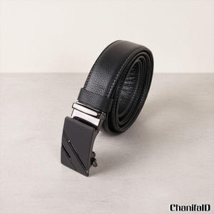 Op de afbeelding: Een zwarte leren riem met een zilveren gesp en een zwarte metalen sluiting. De riem is opgerold, wat de textuur van het leer en de stiksels laat zien. De gesp heeft een modern, hoekig ontwerp.