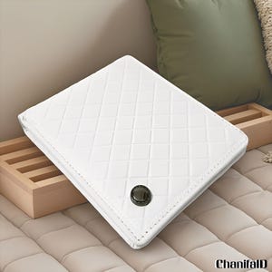 Puede incluir: Una cartera cuadrada blanca con un patrón de diamantes acolchados. La cartera tiene un cierre de botón circular oscuro y está hecha de un material liso. La cartera está sobre una superficie beige.