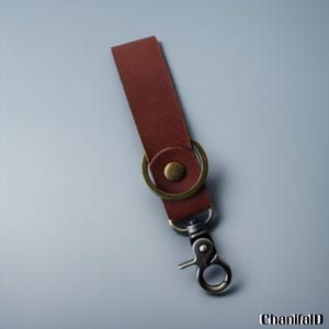 Könnte beinhalten: Ein brauner Leder-Schlüsselanhängerriemen mit einem messingfarbenen Metallring und einem silberfarbenen Metallverschluss. Der Riemen ist rechteckig und hat einen Knopf, der mit dem Ring verbunden ist. Der Verschluss ist vom Dreh-Typ.