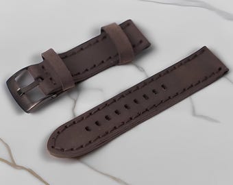 May include: A brown watch strap with a brushed metal buckle and two keepers. The strap is made of a suede-like material and has stitched edges. The watch band is shown on a white marble surface.