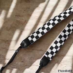 May include: A black and white checkered camera strap. The strap has black leather ends and a black adjustable strap. The checkered pattern is made up of black and white squares. The strap is lying on a wooden surface.