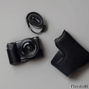 Può includere: Una fotocamera Sony nera con obiettivo, custodia in pelle e cinturino è esposta su una superficie grigia. La fotocamera è in una custodia in pelle nera. Il cinturino è nero e arrotolato. L'obiettivo della fotocamera è visibile.
