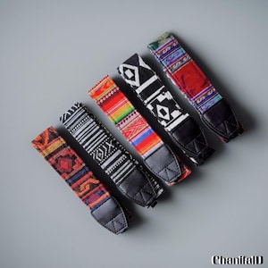 May include: A selection of patterned camera straps. Each strap has a different woven design, with black leather ends. The straps showcase geometric and tribal patterns in red, black, white, and other bright colours. The straps are approximately 80 cm long.