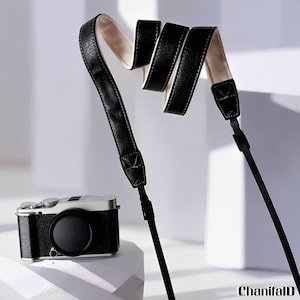 May include: Black leather camera strap with a cream-colored interior, displayed in a wavy pattern. A silver and black camera with a lens cap is positioned below the strap. The strap is attached to the camera with a black strap.