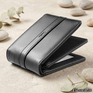 May include: A black leather wallet with a bi-fold design. The wallet has a smooth texture and a strap detail across the front. The brand name "YADA" is subtly embossed on the front. The wallet is open, revealing a clear ID window.