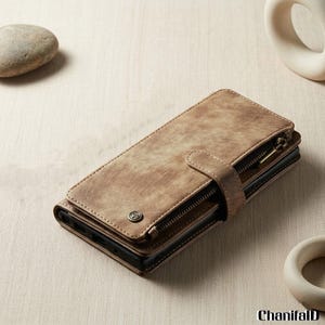 Op de afbeelding: Een bruine leren telefoonhoes met een ritssluiting en een drukknoopsluiting. De hoes heeft een distressed afwerking en een klein metalen embleem. De hoes is ontworpen om een telefoon te bevatten en heeft een portemonnee-achtig ontwerp.