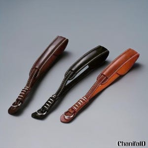 Puede incluir: Tres correas de cuero para cámara en marrón, negro y beige, cada una con costuras blancas y un anillo metálico. Las correas están dispuestas en diagonal sobre un fondo gris, mostrando su diseño y artesanía.