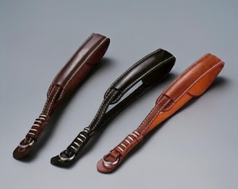 Puede incluir: Tres correas de cuero para cámara en marrón, negro y beige, cada una con costuras blancas y un anillo metálico. Las correas están dispuestas en diagonal sobre un fondo gris, mostrando su diseño y artesanía.