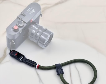 May include: An olive green and black camera wrist strap with a red, white, and blue accent. The strap is attached to a grey camera. The strap is made of a woven material and has a leather accent.