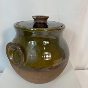 May include: A handcrafted ceramic jar with a lid. The jar has a glossy, two-tone finish, with a dark olive green upper section and a brown base. A circular handle is attached to the side. The lid has a small, brown knob.
