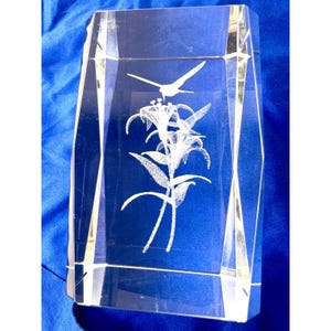 May include: A rectangular clear glass paperweight featuring a three-dimensional laser-etched design of a hummingbird hovering above a cluster of lily flowers. The glass block has bevelled edges and a smooth, polished surface finish.
