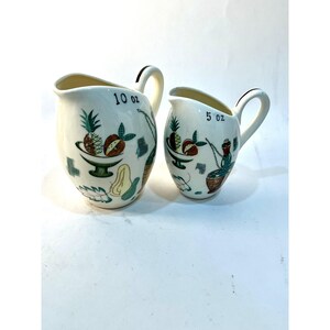 May include: Two cream-coloured ceramic jugs with handles, featuring colourful fruit illustrations and black text. The larger jug is marked 10 oz, and the smaller jug is marked 5 oz. Both vessels have a curved spout and a glossy finish.