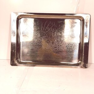 May include: A rectangular, silver-coloured metal tray with an etched landscape design. The design shows a rising sun over mountains and trees. The tray has a slightly raised edge.