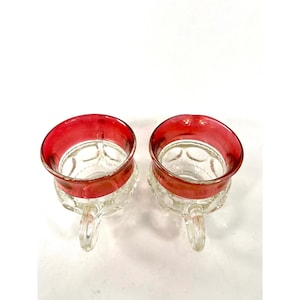 May include: Two clear glass cups featuring a distinct red rim and a textured, circular pattern on the exterior surface. Each cup includes a small, curved glass handle attached to the side, providing a functional grip for these drinking vessels.