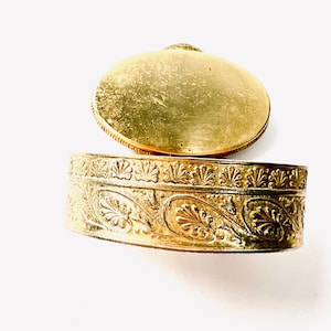 May include: A gold-coloured, oval trinket box with an open lid. The box and lid feature detailed floral and leaf patterns. The box is likely metal and has a vintage or antique appearance, with a slightly worn finish.