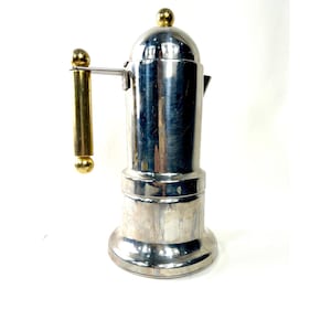 Puede incluir: Una cafetera cilíndrica alta de metal plateado con acabado pulido. Tiene un asa vertical de color dorado unida al lateral, una tapa redondeada rematada con una pequeña esfera dorada y una base ancha y estable para mayor soporte.