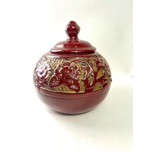 May include: A round, deep red ceramic jar with a matching lid featuring a knob handle. The upper portion of the jar is decorated with a raised, textured floral relief pattern in a contrasting tan colour, while the lower half remains smooth.