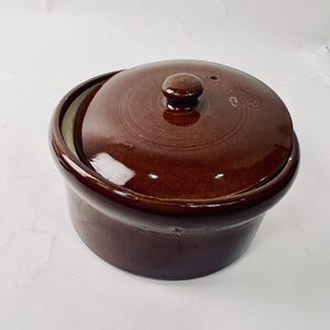 May include: A brown ceramic casserole dish with a lid. The dish is round with a slightly flared rim and a matching lid with a small knob handle. The dish has a glossy finish and appears to be empty.