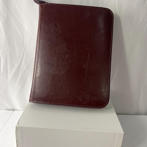 May include: A burgundy leather portfolio with a zipper closure. The rectangular case has a smooth, slightly worn surface. The zipper pull is a darker shade of burgundy. The portfolio is resting on a white box.