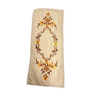 May include: A rectangular cream-coloured table runner featuring intricate embroidery in shades of yellow, gold, and brown. The design includes floral motifs and scrolling patterns arranged in a central oval shape, finished with a scalloped lace edge.