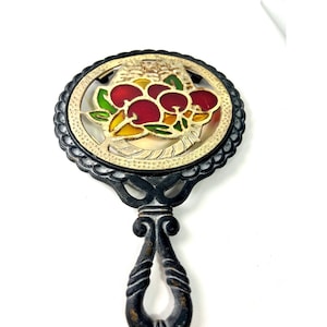Puede incluir: Trivet vintage con mango y marco negros. El diseño circular presenta un anillo interior dorado y un motivo decorativo de cerezas rojas con hojas verdes y amarillas. Un artículo de cocina antiguo.