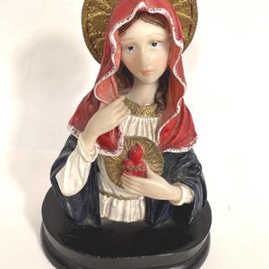 May include: A religious figurine depicting a woman with a red hooded cloak and a gold halo. She wears a white and gold garment and holds a golden heart with a red flame. The figurine is set on a black base.
