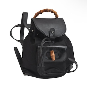 May include: Black backpack with a bamboo handle and accents. The bag features a flap closure and a front pocket with a bamboo clasp. The straps are black leather. The bag is made of a black fabric.