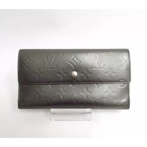 May include: A dark gray leather wallet with a flap closure and a silver snap. The wallet features embossed floral and logo patterns. The wallet is rectangular and appears to be made of high-quality leather.