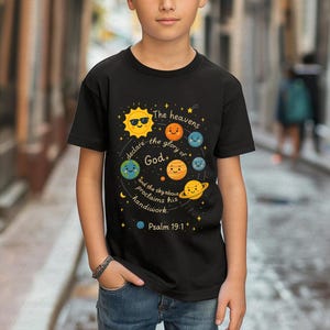 以下が含まれることがあります： 太陽と笑顔の惑星のカラフルなグラフィックが特徴の黒いTシャツ。テキストは「天は神の栄光を語り、大空は御手の業を告げ知らせる。詩篇19:1」と書かれています。