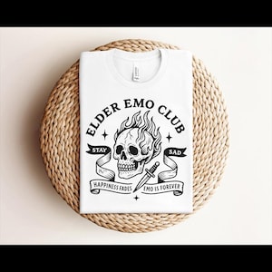Può includere: Maglietta bianca con grafica nera. Il design presenta un teschio con fiamme, un pugnale e le parole "ELDER EMO CLUB". Gli striscioni recitano "STAY", "SAD", "HAPPINESS FADES" e "EMO IS FOREVER".