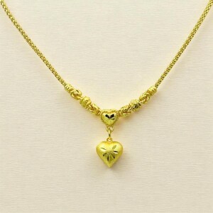 May include: A gold-colored necklace with a heart pendant. The necklace features a delicate chain and decorative beads. The heart pendant has a textured surface with a floral design. The necklace is displayed against a neutral background.