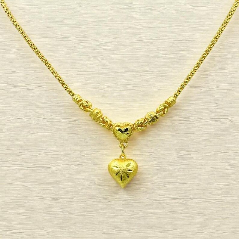 May include: A gold-colored necklace with a heart pendant. The necklace features a delicate chain and decorative beads. The heart pendant has a textured surface with a floral design. The necklace is displayed against a neutral background.