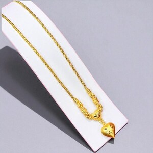 May include: A gold-colored necklace with a heart-shaped pendant. The necklace features a delicate chain and a heart pendant with a cut-out design. The necklace is displayed on a white surface.