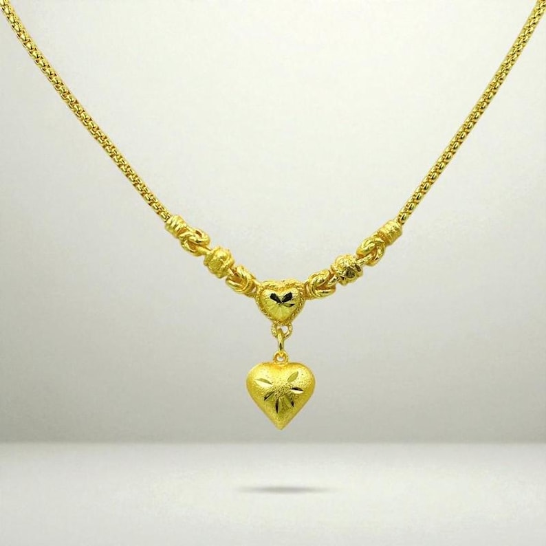 May include: A gold necklace featuring a heart pendant. The necklace has a delicate chain and decorative elements near the pendant. The heart pendant has a textured surface with a starburst design.