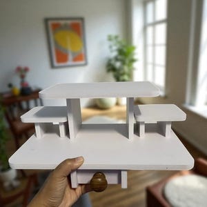 Può includere: Un espositore bianco a più livelli con una base rettangolare, un livello intermedio e due piattaforme laterali. L'espositore ha un pomello di legno sul fondo. L'espositore è tenuto da una mano.