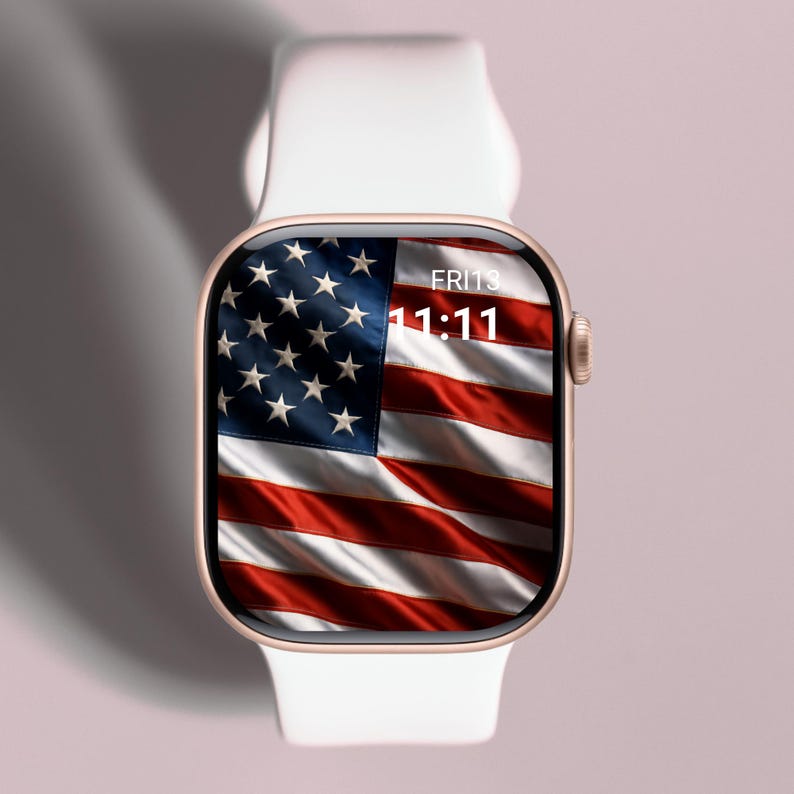 May include: A gold-colored smartwatch with a white band displays the American flag on its screen. The time is 11:11 on Friday. The watch is set against a soft pink background.