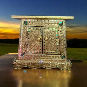 May include: A gold-colored, ornate cabinet with two doors, embellished with green and iridescent accents. The cabinet has a rectangular shape with a flat top and is set against a sunset backdrop.