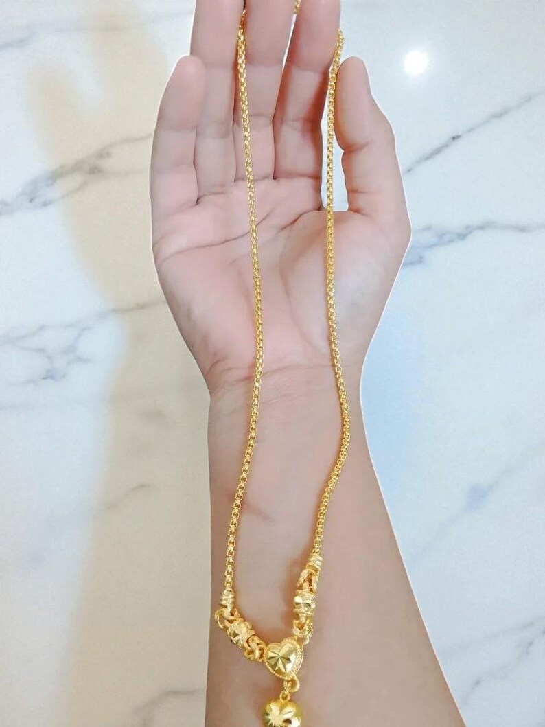 May include: A gold-colored necklace with a heart-shaped pendant and a small, round charm. The necklace is held in a hand, showcasing its intricate design and delicate chain. The pendant has a textured surface.