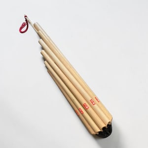 May include: A light-colored, wooden instrument with multiple pipes, likely a wind instrument. The pipes are arranged in a fan shape and have red markings. A red string is attached to the top. The instrument has a black base.