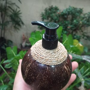 Può includere: Un dispenser di sapone sferico marrone scuro realizzato con un guscio di cocco. Il dispenser ha una pompa nera ed è adornato con un dettaglio in corda intrecciata attorno al collo. Lo sfondo presenta un rigoglioso fogliame verde.