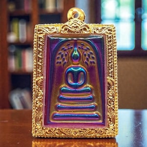 Puede incluir: Colgante rectangular dorado con un marco detallado. El colgante presenta una figura de Buda sentado en tonos de azul, morado y dorado. El fondo tiene un efecto degradado. El colgante tiene un lazo en la parte superior para una cadena.