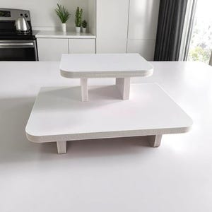 May include: Two-tiered white serving stands. The larger stand is rectangular, and the smaller one is square. Both have a white surface and a textured, light gray edge. The stands are elevated on short, white legs, suitable for displaying food or decor.