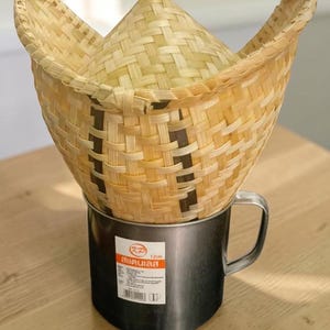 May include: A woven bamboo steamer basket with a conical lid, set atop a stainless steel pot with a handle. The basket is light brown, and the pot has a label with text.