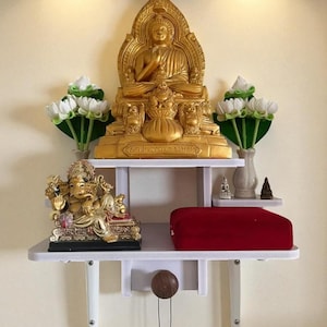 Può includere: Una mensola bianca a parete con una statua dorata di Buddha, una statua dorata di Ganesha e un cuscino da meditazione rosso. Fiori di loto bianchi e verdi decorativi sono disposti su entrambi i lati della statua del Buddha.